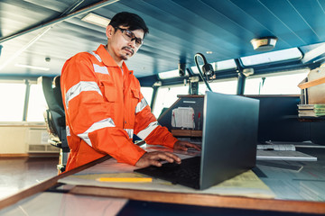 Filipino deck Officer on bridge of vessel or ship wearing coverall during navigaton watch at sea . He is using laptop, electronic paperwork at sea, concept of reporting