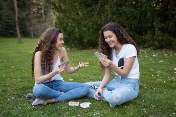 Twins sisters playing cards laying in the grass in a public park.