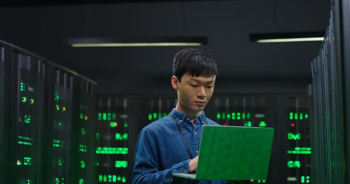 Male Asian system administrator of big data center typing on laptop computer while working in server room. Programming digital operation. Man engineer working online in database center. Programming.