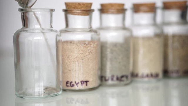 Pouring sand from vacation into small transparent jar standing next to already full ones with names of different countries on them.