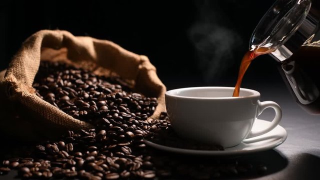 Pouring coffee with smoke on a cup and coffee beans on burlap sack on black background.