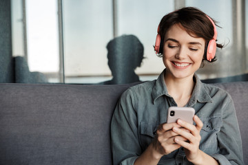 Image of nice young smiling woman using smartphone and headphones - obrazy, fototapety, plakaty