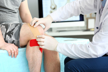 Close-up of hurt knee bound with red health care and protective, sticky red bandages. Doctor helping patient putting health care patch on injured, aching leg. Medical treatment concept