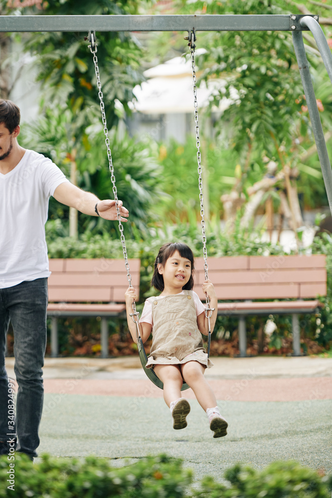 Wall mural Father pushing his adorable little daughter sitting on swing in park