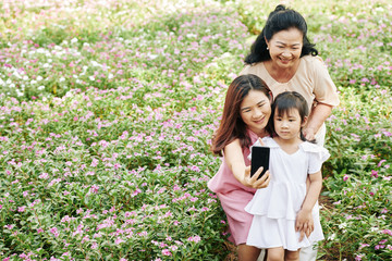 Smiling pretty Asian woman taking selfie with her senior mother and little daughter outdoors