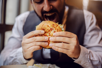 Closeup of hungry man in suit sitting in fast food restaurant and eating cheese burger.