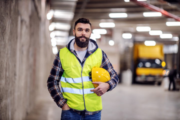 Cute bearded construction worker in vest and with safety helmet in hands standing inside of building he renovating.