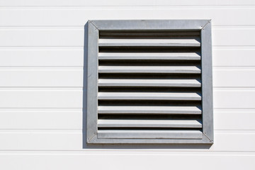 Ventilation hole with stainless steel grill. Wall made of light plastic siding. Horizontal arrangement of the stripes.