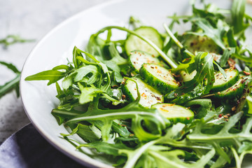 Green salad of arugula and cucumbers with oil and spices in gray dish.