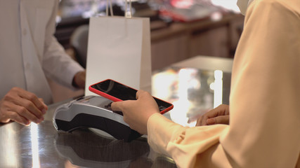 An upwardly mobile Asian Muslim woman using a mobile phone - smartwatch to pay for a product at a sale terminal with nfc identification payment for verification and authentication