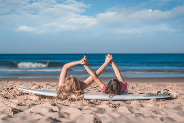 Two little girls' hands holding together laying on a surfboard on the sandy ocean beach. Love, freindship, togetherness concept.