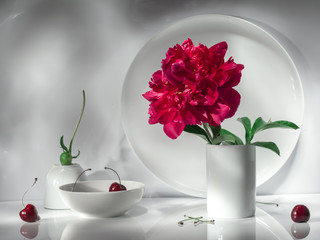 Still life in a minimalist white color scheme. One white peony flower in a vase against a white circle, 2 bowls with a peony leaf.