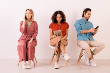 Modern multi-ethnic people in casual outfits sitting on chairs in row and using gadgets all time for communication