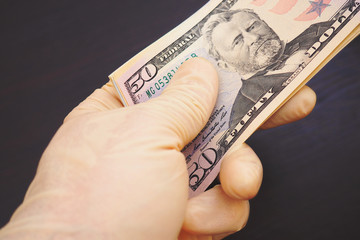 A hand in a flesh-colored latex glove holds a bundle of American US money. Above is a 50 dollar bill. Dark brown background. Precautions, prophylaxis against coronavirus, COVID-2019 during a pandemic