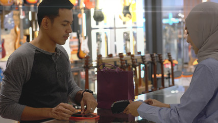 An upwardly mobile Asian Muslim man using a mobile phone - smartwatch to pay for a product at a sale terminal with nfc identification payment for verification and authentication