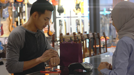 An upwardly mobile Asian Muslim man using a mobile phone - smartwatch to pay for a product at a sale terminal with nfc identification payment for verification and authentication