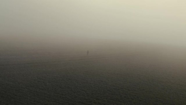 A drone following a man walking on a field through thick fog. A scene that looks like it is from a Blade Runner Movie.