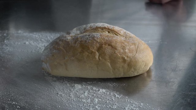 A baker sprinkles flour using his hand over a bread boule on the bakery's stainless steel table, slow motion close up shot.