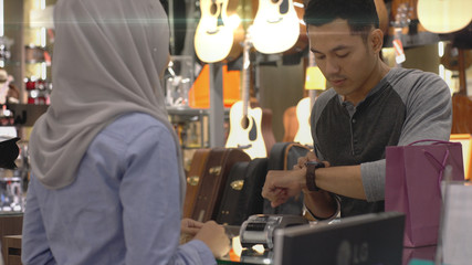 An upwardly mobile Asian Muslim man using a mobile phone - smartwatch to pay for a product at a sale terminal with nfc identification payment for verification and authentication