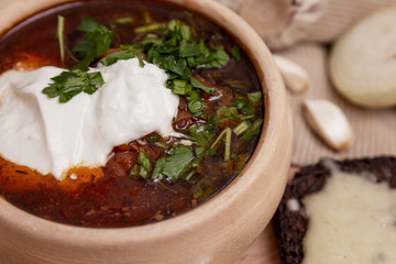 Appetizing Russian borsch with sour cream and green pepper in a clay bowl on a wooden board. Served with garlic and onions. A popular dish of national cuisine.