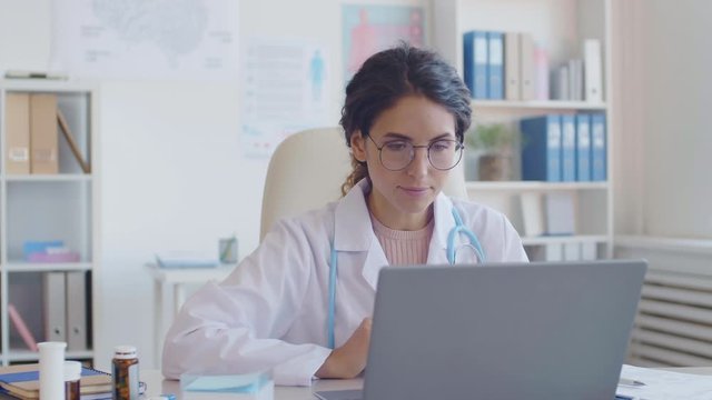 Panning of young female doctor wearing medical gown and stethoscope sitting at desktop in front of laptop and starting conversation with patient via internet