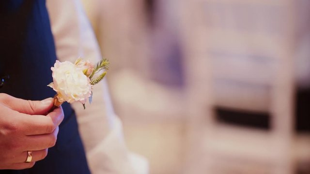 The caucasian groom in a black vest with a gold wedding ring holds a buttonhole in his hand