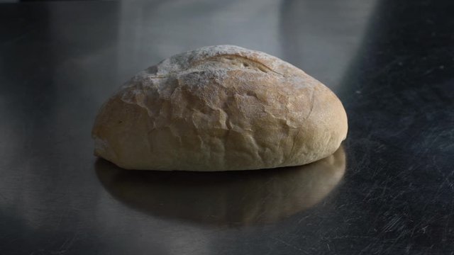 A baker sprinkles flour using his hand over a bread boule on the bakery's stainless steel table, slow motion close up shot on different perspective