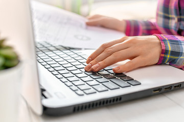 Young woman works on laptop at home. Hands holding a sheet of document and typing on a keyboard closeup. Remote work concept, freelance, quarantine, study.