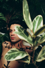 A young handsome brunette in green foliage. Caucasian man with body art near the ficus
