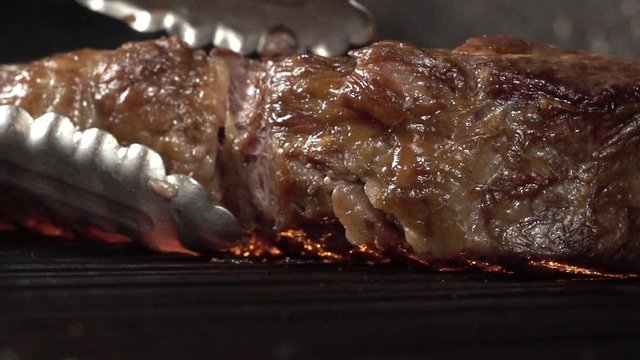 A tasty tomahawk steak on the cooking grill fire, extreme close up slow motion shot. Using a tong to turning the piece of meat over the grilling flame.