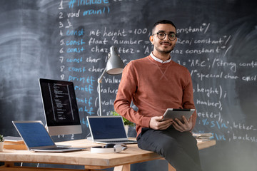 Portrait of confident young Arabian coder in glasses sitting on table and using tablet against...