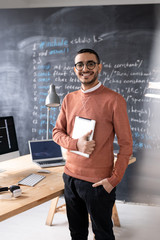Portrait of cheerful young middle-eastern IT specialist in glasses standing with tablet in modern office, computer language script on blackboard