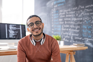 Portrait of smiling young Arabian programmer in eyeglasses sitting in modern office of IT company