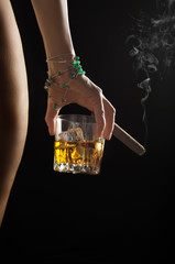 Caucasian model holds glass of whiskey with icecubes and smoking cigar in her hand near naked buttock and hip on dark background closeup studio shot - obrazy, fototapety, plakaty