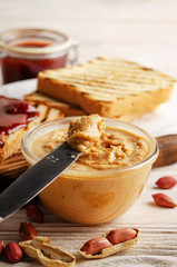Low angle view at glass bowl with peanut butter on white wooden table with toasts and jam aside....