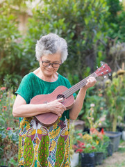 Portrait of elderly woman playing ukulele in garden. Relaxing by singing and play small guitar happy and enjoy life after retired. Concept of old people and health care. Space for text