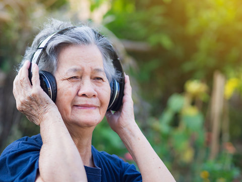 Elderly woman standing wearing wireless headphone listening to a favorite song, and smile happy in garden. Asian senior woman, short white hair are enjoying the music. Good mental health concept