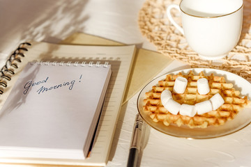 Marshmallow waffle smile greeting card good morning on white textured table. Flat composition.