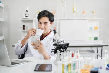 A male scientist with black hair wearing white coat using a mobile phone device sipping coffee with...