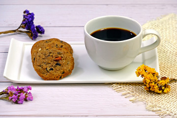Homemade soft and chewy chocolate chip cookies and a cup of black coffee decorated with dried flowers on white wooden table.