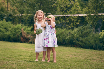 Two little girls in sarafans and pigtails in a green garden are holding broccoli in their hands. They close their eyes, laugh. Healthy food concept, green vegetarian food.