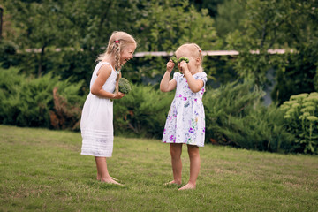 Fototapeta premium Two little girls in sarafans and pigtails in a green garden are holding broccoli in their hands. They close their eyes, laugh. Healthy food concept, green vegetarian food.