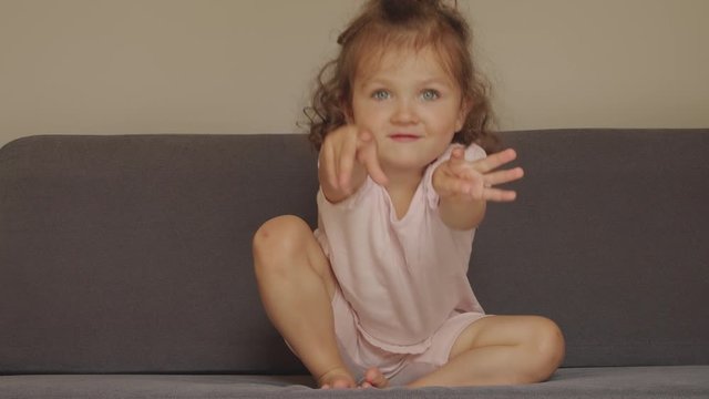 Charming funny little girl sitting on a sofa asks to pick her up. Baby hugs.