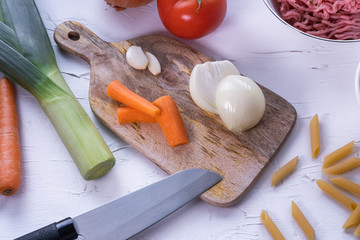 Onion, carrot and cloves of garlic on a wooden table with other ingredients to prepare pasta bolognesa on white textured background. Italian cuisine. Top view.