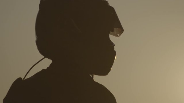 Hand held slow motion shot of male silhouette wearing and adjusting a car racing helmet with sun setting in background
