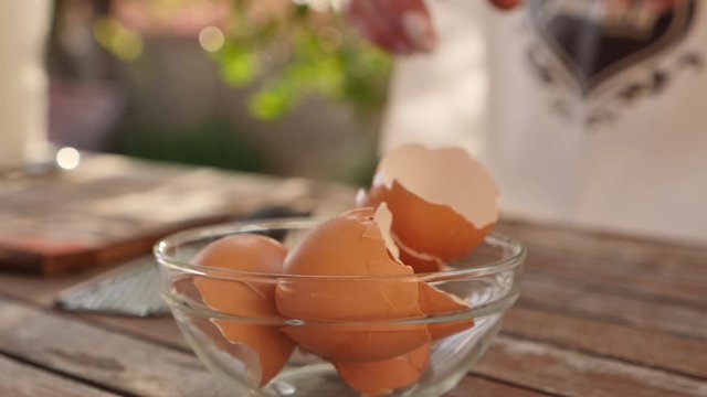 woman putting remaining egg shell into a glass bowl full of egg shells in a wooden table
