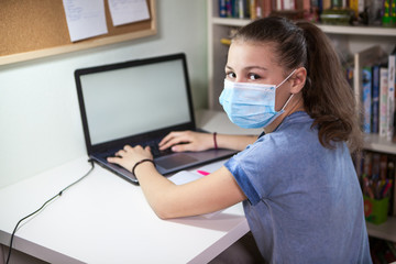 Preteen Caucasian girl in medical mask using her laptop, printing on keyboard, schoolchild studying at home during covid-19 pandemic