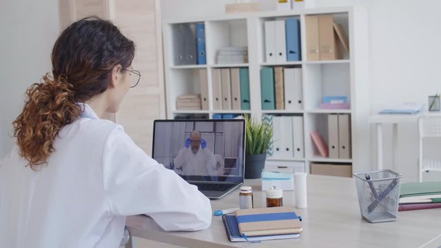 Back view of curly-haired female doctor wearing medical gown sitting at desk in front of laptop and communicating with her male colleague distantly