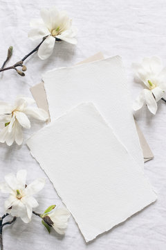 Wedding stationery set. Mock-up scene with blank paper greeting cards, envelope on linen tablecloth background. White magnolia stellata tree branches. Feminine still life, flat lay, top view.