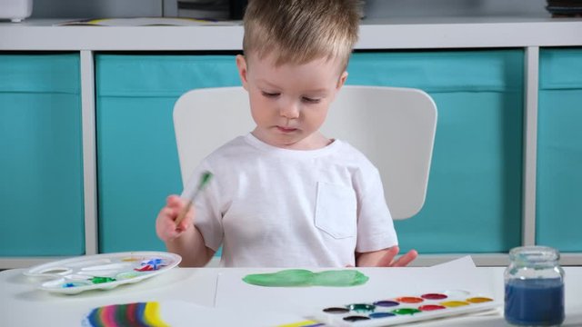 Blonde Caucasian boy artist in children's room draws green grass at table with watercolors, child wetting the brush in water, applying paint to brush and drawing on paper
